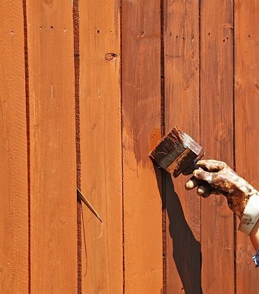 Hand holding a brush applying dark creosote preservative to an orange-stained wooden fence